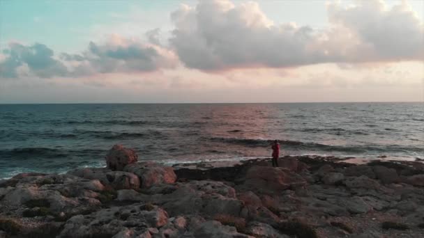 Charming caucasian girl standing on big stone at rocky seashore and taking photos and videos of beautiful mediterranean sea at sunset. Drone aerial shot — Stock Video