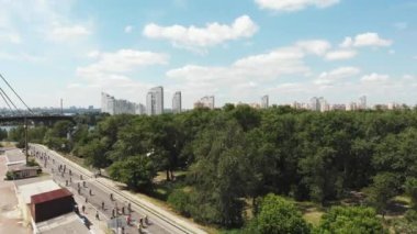 Parlak aktif giysiler içinde genç çekici erkek ve bayan bisiklet sürücüleri parkın turistik bölgesi boyunca güzel ağaçları ve ufukta büyük beton binaları olan yolda ilerliyorlar. Hava aracı görüntüsü