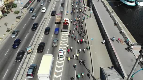 Vue aérienne de l'autoroute de la ville avec embouteillage et groupe de jeunes cyclistes heureux en vêtements de sport lumineux chevauchant le long du beau quai avec des bateaux lors d'une journée chaude ensoleillée — Video