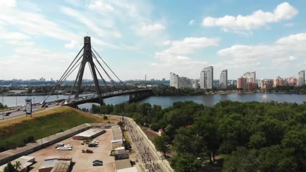 Gruppe junger attraktiver Radfahrer, die durch Park mit großen grünen Bäumen und Erholungsgebiet in der Nähe des Flusses Dnipro fahren. Drohnenaufnahmen von der Moskauer Brücke und Radfahrern, die die Parkpromenade überqueren, mit dem großen Stadtzentrum am Horizont — Stockvideo