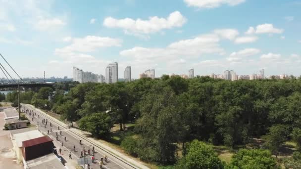 Jovens atrativos ciclistas masculinos e femininos em trajes ativos brilhantes andando na estrada ao longo da área turística do parque com belas árvores e grandes edifícios de concreto no horizonte. Drone aéreo disparado — Vídeo de Stock