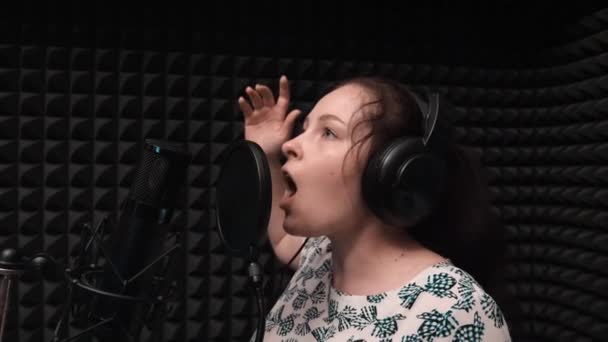 Ένα νεαρό κορίτσι με ακουστικά που κάνουν πρόβες στο μουσικό στούντιο φωνητικής. Ελκυστική γυναίκα καταγράφει το τραγούδι — Αρχείο Βίντεο