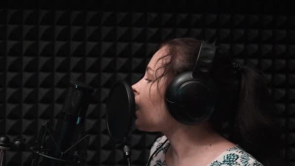 Menina atraente em fones de ouvido cantando em estúdio vocal profissional com fundo preto — Vídeo de Stock