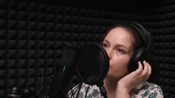 Close up de menina morena jovem com olhos azuis. Mulher atraente em fones de ouvido cantando ao microfone no estúdio — Vídeo de Stock