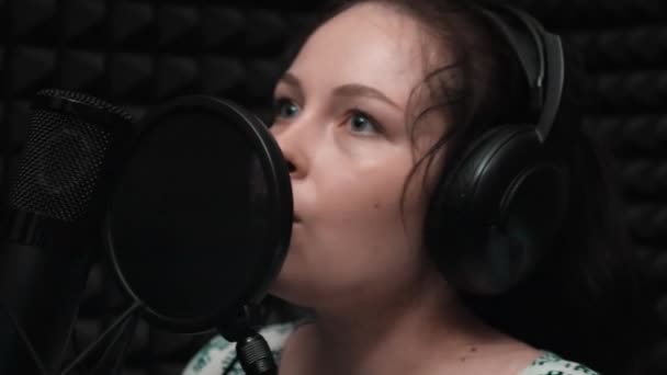 Close up side view of face of attractive brunette girl is recording voice to microphone in music studio with black background. Close up view of microphone and music equipment at recording studio — Stock Video