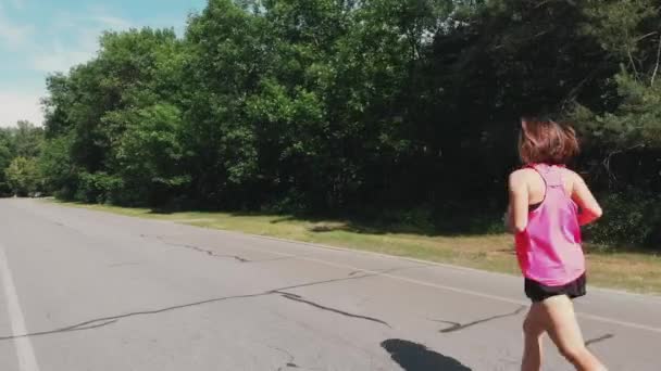 Junge attraktive Läuferin. schöne motivierte Mädchen in rosa Hemd läuft im Stadtpark. Sportliche Frau mit Kopfhörer trainiert im Park — Stockvideo