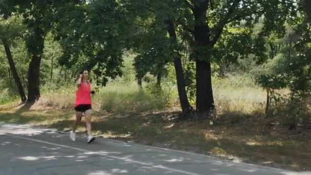 Ung sportiga flicka i rosa skjorta och korta svarta shorts som körs i parken. Attraktiv kvinna i hörlurar med utbildad kropp gör övningar i skogen. Brunett kvinna som kör i parken. Running Girl — Stockvideo