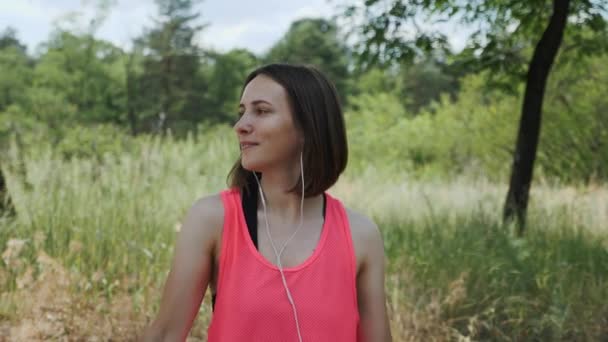 Chica deportiva atractiva en la parte superior rosa con auriculares escuchando música en el teléfono inteligente. Chica morena delgada bailando en el parque. Chica joven sonriendo a la cámara. Concepto de funcionamiento. Movimiento lento — Vídeo de stock