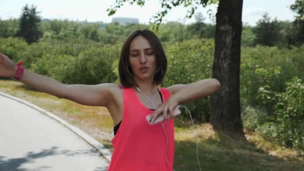 Menina atraente quente nova dançando no parque. Menina esportiva na camisa rosa sorrindo. Menina morena em fones de ouvido com smartphone olhando para a câmera e sorrindo. Menina bonito está se divertindo no parque. Movimento lento — Vídeo de Stock