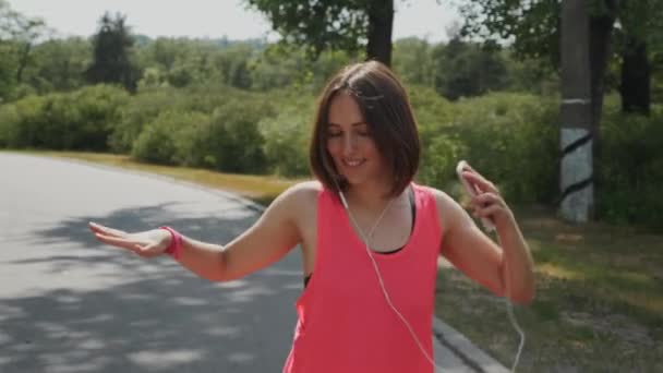 Attraente ragazza sportiva godendo la sua passeggiata nel parco. Giovane donna in cuffia che balla. Ragazza carina in camicia rosa sorridente. Bella ragazza caucasica che ascolta musica. Rallentatore — Video Stock