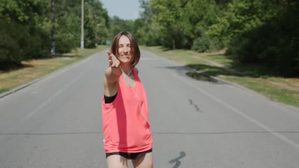 Attractive girl in headphones looking to camera in park. Sportive woman in pink shirt smiling and having fun in park. Caucasian brunette girl inviting to follow her. Girl in sportswear — Stock Video