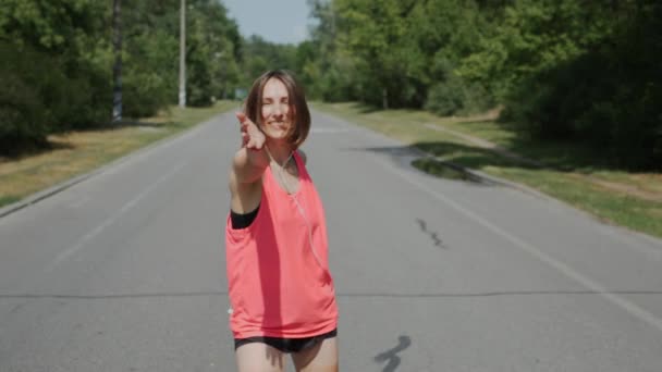 Attractive girl in headphones looking to camera in park. Sportive woman in pink shirt smiling and having fun in park. Caucasian brunette girl inviting to follow her. Girl in sportswear. Slow motion — Stock Video