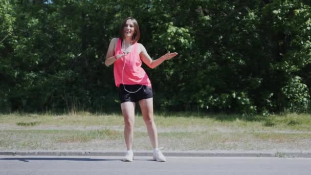 Přitažlivá Kavkazská dívka v růžové košili se sluchátky v parku. Mladá žena dělá směšné a směšné pohyby. Dívka se usmívá a baví se v parku. Zpomaleně — Stock video