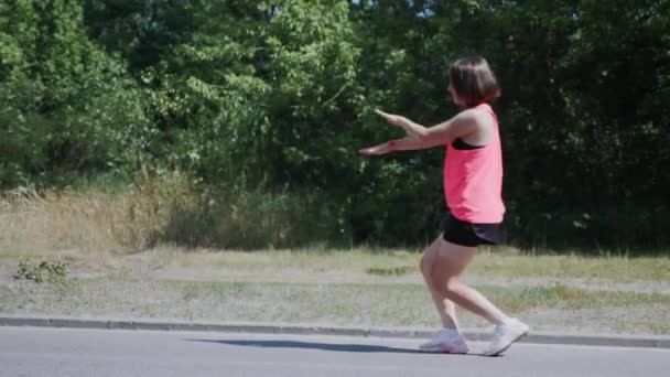 Söt brunett tjej dansar i parken. Attraktiv ung kvinna lurar runt och ha kul. Flicka i hörlurar utför roliga drag. Söt flicka i rosa skjorta i parken — Stockvideo