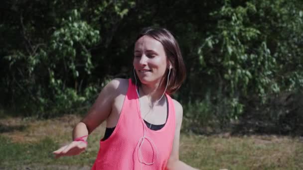 Menina feliz encantadora em top rosa e fones de ouvido olhando e sorrindo para a câmera. Mulher quente atraente dançando e ouvindo música no parque. Menina cantando em fones de ouvido. Menina bonita apreciando música — Vídeo de Stock