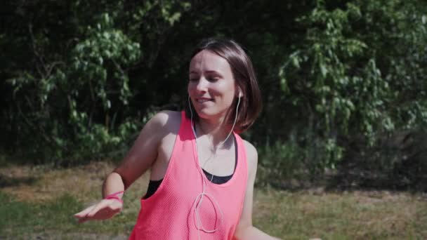 Affascinante ragazza felice in top rosa e cuffie guardando e sorridendo alla fotocamera. Attraente donna calda ballare e ascoltare musica nel parco. Giovane ragazza che canta. Bella ragazza che ama la musica. Rallentatore — Video Stock