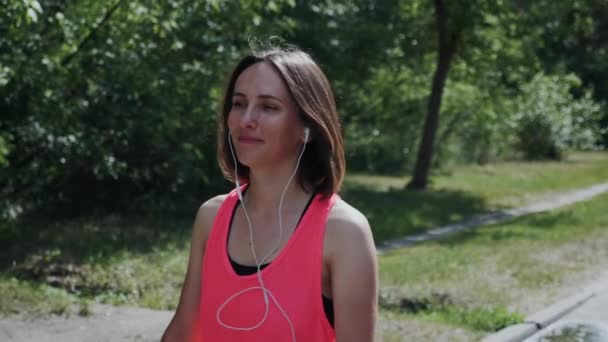 Young happy girl in headphones walking in park. Attractive woman received message to smartphone. Girl looking to phone and writing text message in park. Girl listening music in park. Slow motion — Stock Video