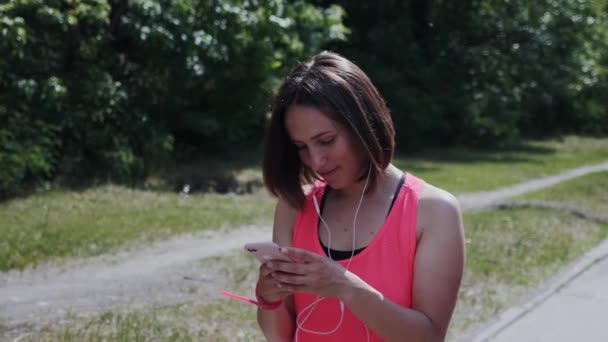 Söt flicka i hörlurar går genom parken. Attraktiv ung kvinna i hörlurar är textning meddelande. Flicka tittar på smartphone och leende. Flickan fick ett textmeddelande och upprörd. Slow motion — Stockvideo
