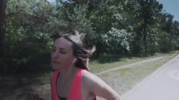 Muchacha deportiva atractiva en camisa rosa corriendo en el bosque. Mujer delgada con cuerpo entrenado haciendo ejercicios. Chica en los auriculares está respirando fuertemente durante el entrenamiento. Concepto de funcionamiento. Movimiento lento — Vídeos de Stock