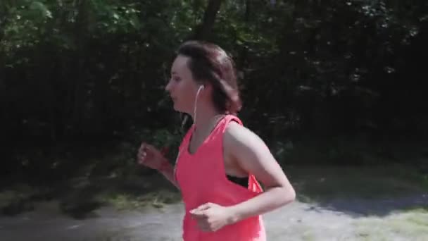 Chica delgada atractiva en la parte superior de color rosa brillante con auriculares está respirando pesadamente durante el entrenamiento. Una joven deportista corriendo en el parque. Entrenamiento de chicas en el parque. Mujer haciendo ejercicios de correr — Vídeos de Stock