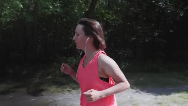 Attraktiv smal flicka i ljust rosa topp med hörlurar andas tungt under träningen. Ung sportiga kvinna som springer i parken. Flicka utbildning i stadsparken. Kvinna gör övningar. Slow motion — Stockvideo