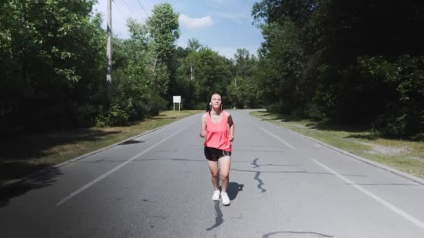 Menina magro nova na camisa rosa com fones de ouvido está começando a correr no parque. Morena mulher atraente com corpo treinado se preparando para corrida. Menina com bronzeado ciclismo correndo no parque. Movimento lento — Vídeo de Stock