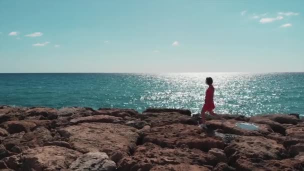 Vrouw in rode jurk wandelen op rotsachtige Pier strand met golven raken kliffen en zon schijnt op zonnige dag. Sun Road reflectie op blauw water en hemel. Meisje in rode wandelingen op rotsen langs zee in Cyprus — Stockvideo