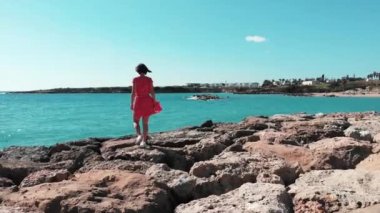 Kıbrıs'ta Mercan Körfezi'ndeki kayalık kayalık larda yürüyen kırmızı ve beyaz elbiseli kadın. Kayalık plajda kırmızı giyinmiş kadın havadan görünümü. Sadece kayalar üzerinde kırmızı elbiseli kız Drone atış.