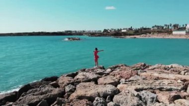 Güneş yolu ve mavi deniz suyu bakarak kayalık kayalık üzerinde ayakta sarkan rüzgar ile kırmızı sevimli elbise havada elleri ayrı kadın. Güneşin tadını çıkarırken havada kollarını ayrı kırmızı bayan Havadan görünümü