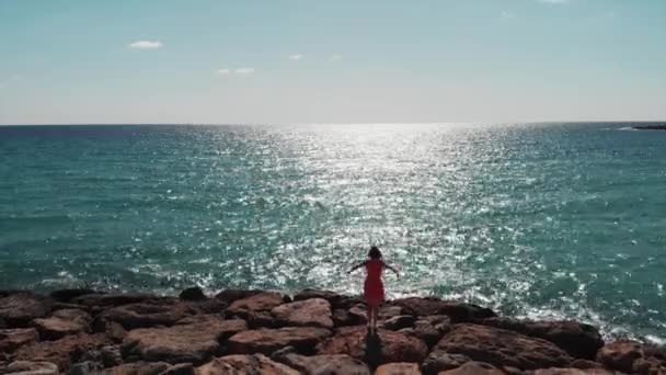 Silhueta escura de mulher em vestido vermelho com braços separados no ar em pé no cais de praia rochosa contra o sol. Sol reflexão estrada na água azul com céu e vento soprando. Vista aérea da menina com as mãos afastadas — Vídeo de Stock