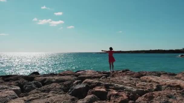 Elleri kayalık kayalıklarda duran kırmızı elbiseli bayan güneşli bir günde mavi denize bakıyor. Kırmızı elbise rüzgar ve güneş üzerinde su yansıtan çırpan. Elleri ayrı olan kadının havadan drone görünümü — Stok video