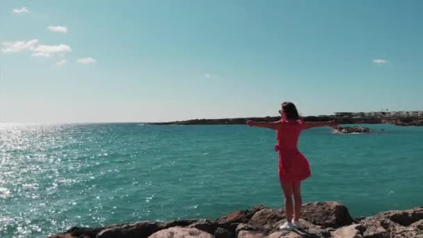 Dame in rood flappen op wind jurk staande op rotsachtige strand Cliff Pier met handen uit elkaar op zonnige zomerdag op Cyprus Coral Bay. Aeral weergave van de vrouw in rode outfit op rotsachtige strand met armen uit elkaar. — Stockvideo