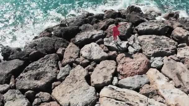 Kobieta w czerwonej pięknej sukni z rąk od siebie w powietrzu stoi na skalistym klifie plaży z fal morskich zderza się z kamieniami rozpryskiwania wody wszędzie i białej pianki. Cypr Coral Bay aerial widok z góry — Wideo stockowe