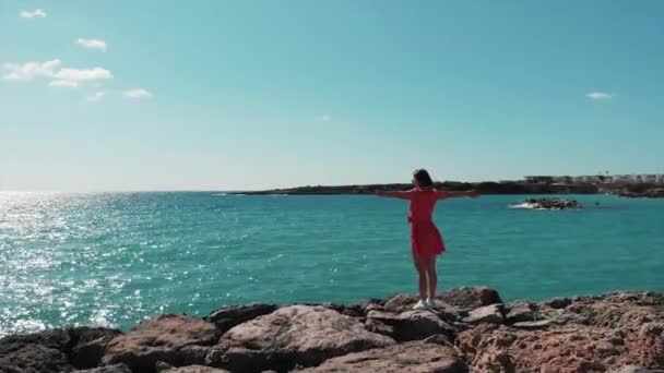 Woman in red dress with hands apart against sun standing on rocky cliff pier enjoying wind and sun. Ocean waves hitting rocky beach splashing and creating white foam. Aerial drone shot. — Stock Video