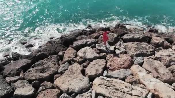身着红色礼服的女士站在岩石码头或悬崖上，双手分开在空中，看着蓝色的大海和天空，阳光反射在水面上。珊瑚湾在塞浦路斯帕福斯鸟瞰图 — 图库视频影像