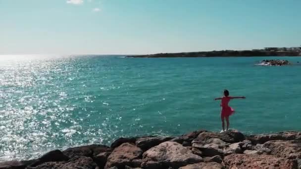 Donna in abito rosso con le mani divaricate in aria in piedi sulla spiaggia rocciosa scogliera con drone che vola intorno a lei. Veduta aerea della signora silhouette scura in abito rosso e bianco con le mani divaricate in piedi su rocce — Video Stock