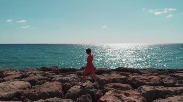 Güzel kadın siluet kayalık deniz iskelesi üzerinde tek başına yürüyor. Deniz dalgaları kayalık plaja çarpar. Güneşli sıcak bir günde kayalık mercan plaj boyunca yürüyen kırmızı elbiseli kız ile mavi denizde Güneş yol yansıması — Stok video