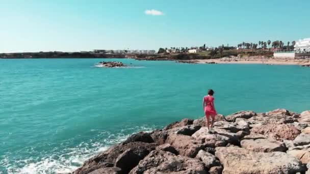 Nő a piros séta a sziklás strandon, és emeli a kezét egymástól a levegőben nézi a kék óceán és ég. Szél csapkodó ruhája. Coral Bay Cyprus és a hölgy piros ruhában, karokkal kívül sziklás strand — Stock videók
