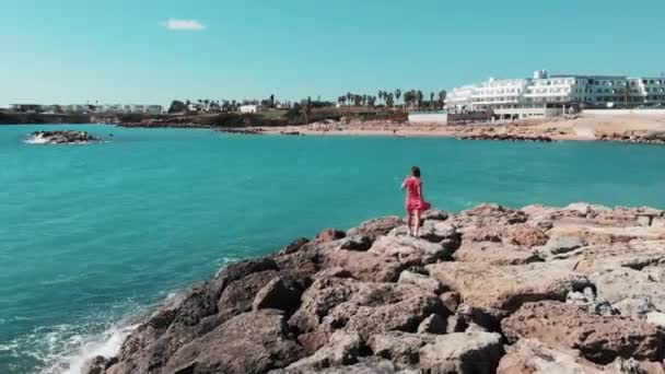 Kvinna i rött på stenig strandklippa. Lady i röd klänning njuter av sol och varm vind på Coral Bay Cypern. Framgångsrik hona i röd och vit klänning promenader längs Rocky Beach Pier i Cypern Paphos — Stockvideo