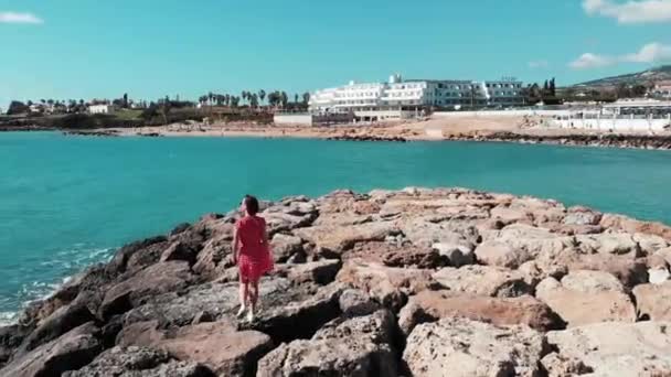 태양과 뜨거운 바람을 즐기는 혼자 바위 해변 부두에 걷는 빨간 드레스에 귀여운 여성. 바위와 태양과 충돌하는 파도는 태양 도로를 만드는 물에 반영됩니다. 공중 무인 항공기 샷 — 비디오