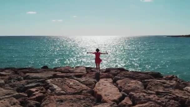 Silueta de Mujer vestida de rojo con los brazos separados en el cielo de pie sobre rocas en el muelle de la bahía de coral mirando al mar y al sol reflejando la carretera en el agua azul. Foto aérea de dama en vestido rojo con las manos separadas — Vídeos de Stock