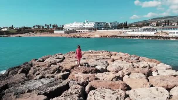 Mujer vestida de rojo hermoso vestido caminando sola en el muelle rocoso en la bahía de coral cyprus con hotel y playa en el fondo. Las olas azules del mar golpean la playa rocosa con espuma blanca y salpicaduras de agua. Disparo aéreo — Vídeos de Stock