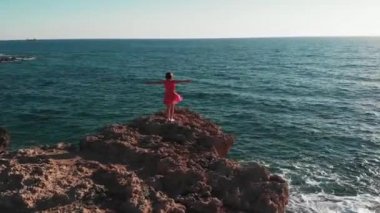 güneş ve güneş yoluna karşı kayalık kayalık üzerinde duran havada ayrı elleri ile kadın siluet. Kırmızı elbiseli kız, kollarını ayırarak rüzgarda çırpınan dağ uçurumunda duruyor. Drone'un havadan çekilmiş kadın fotoğrafı