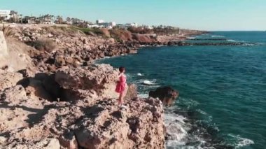 Dağ uçurumunun tepesinde kırmızı elbiseli ve elleri havada. O güneş ve sıcak rüzgar esme tadını çıkarırken Rüzgar elbisesini çırpma. Kolları ayrı ve deniz dalgaları çarpışan kadının Drone görünümü