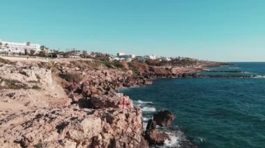 Kayalık kayalıklarda kırmızı elbiseli kadın, elleri havada yanlara. Beyaz güçlü deniz dalgaları deniz kıyısına vurarak beyaz köpük ve sıçrama. Dağ yamacının tepesinde kırmızı elbiseli kadın. Drone atış