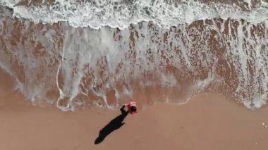 Deniz kıyısında yürüyen bir kız. Sahilde yürüyen kızın havadan üst görünümünü kapatın. Sahil şeridinde kırmızı kıyafetli kadın. Kumda ayak izi. Kadının ayakları kumda yürür. Okyanus sahilinde yürüyen güzel kadın
