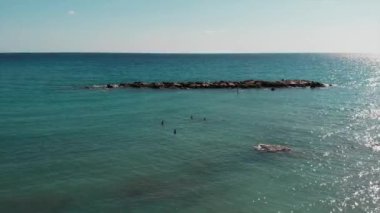 Drone kayalık deniz kıyısına yakın uçuyor. Mavi deniz suyunun havadan görünümü. Güzel deniz manzarası. Sup genç çekici adam. Güneş tabaklanmış çocuk sup üzerinde yüzen. Mutlu erkekler yüzme ve güneşin tadını çıkarıyor