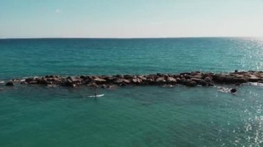 Sup'ta yüzen adam. Genç çocuk sörf yapıyor. Güzel deniz manzarası havadan görünümü. Deniz kayalık taşlarından drone atışı. Mavi berrak suyla denizde yüzen erkekler. Erkekler rahatlatıcı ve güneşin tadını çıkarıyor