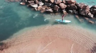 Sörf tahtasında yüzen güneş lekişmiş genç bir adam. Kumlu plaj yakınında sörf sportif çocuk. Supboard'daki çocuğun havadan görünümü. Büyük kayalar ile mavi berrak deniz suyu Drone atış