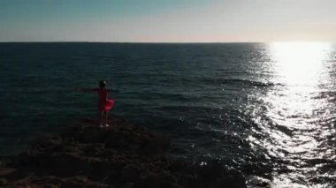 Kayalık sahildeki kız. Okyanus kayalıklarında duran genç bir kadın. Denizde kırmızı elbiseli kız. Gün batımına bakan çekici bir kadın. Güzel okyanus gün batımı. Büyük kayalık taş üzerinde duran kızın havadan drone görünümü
